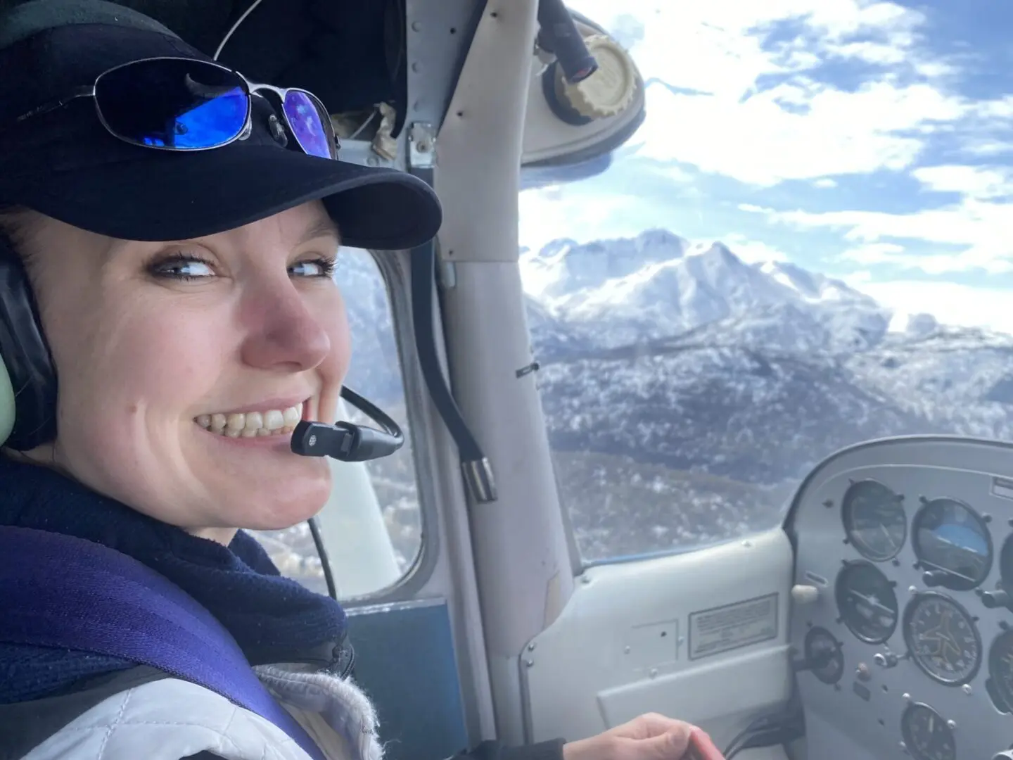 A woman smiling in the cockpit of a plane with mountains in the background.