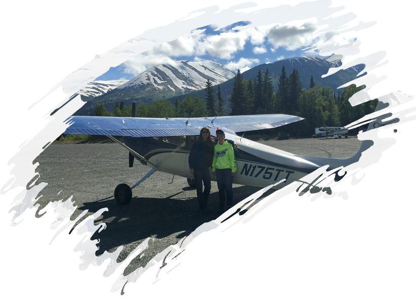 Two people standing in front of a small plane with mountains in the background.