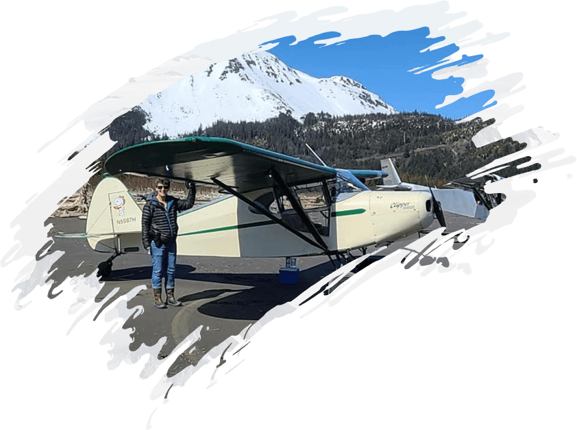 A person standing next to a small plane with mountains in the background.